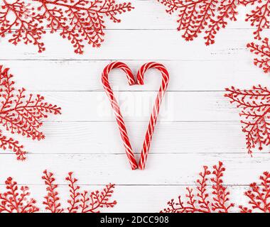 Herzform aus Bonbonstöcken auf weißen Holzplanken mit roten Glitzerzweigen umrahmt. Weihnachtskonzept. Stockfoto