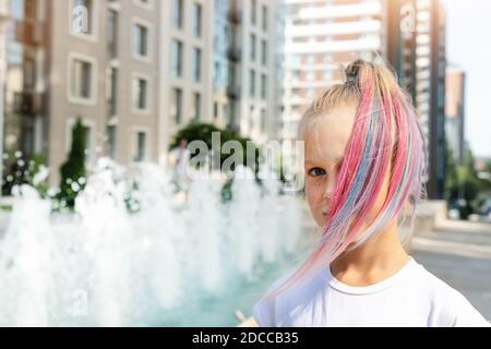 Niedliche liebenswert kaukasischen kleinen Mädchen mit bunten Pastellkreide gefärbt blonde Haare auf der Straße der Stadt mit Brunnen Hintergrund gemalt. Weibliches Kind Stockfoto