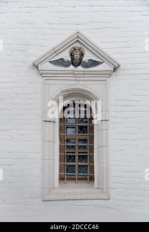 Altes Bogenfenster mit Metallgitter und Relief auf einem Weiße Ziegelmauer der christlichen Kirche Stockfoto