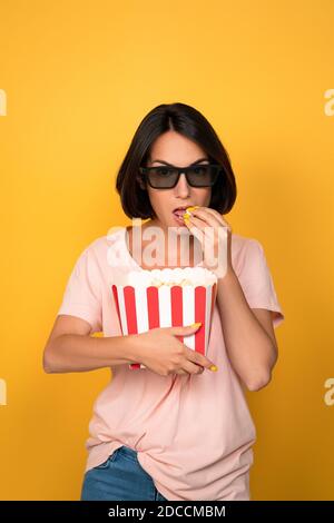 Junge Brünette Frau mit Eimer Popcorn in Stereo-Gläser. Kinokonzept. Isoliert auf gelbem Hintergrund Stockfoto