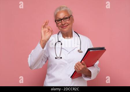 Ältere Ärztin lächelnd und gestikulend ok isoliert auf einem Rosa Hintergrund Stockfoto