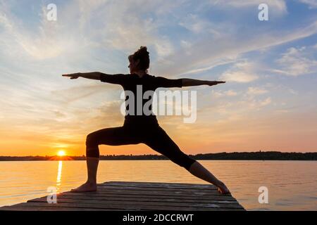 Frau praktiziert Yoga Haltung Virabhadrasana II / Krieger Pose, lunging stehen Asana auf Anlegesteg am See bei Sonnenuntergang Stockfoto