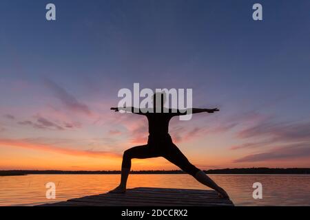 Frau praktiziert Yoga Haltung Virabhadrasana II / Krieger Pose, lunging stehen Asana auf Anlegesteg am See bei Sonnenuntergang Stockfoto