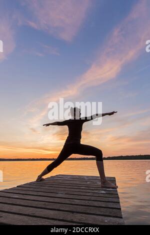 Frau praktiziert Yoga Haltung Virabhadrasana II / Krieger Pose, lunging stehen Asana auf Anlegesteg am See bei Sonnenuntergang Stockfoto