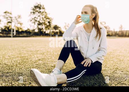 Athletische junge Frau in einer medizinischen Schutzmaske auf ihrem Gesicht, Ruhe nach dem Training auf dem Boden unter dem offenen Himmel sitzen Stockfoto