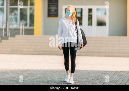 Ein junges Mädchen in einer medizinischen Schutzmaske, mit einer Aktentasche vor dem Hintergrund eines Gebäudes, im Freien stehend. Quarantäne, Coronavirus Stockfoto