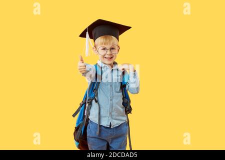 Kind Junge Grundschüler in einer Graduiertenmütze, Brille und mit einem Rucksack auf einem gelben Hintergrund. Zeigt mit dem Finger ein OK-Zeichen. Wissensstand Tag A Stockfoto