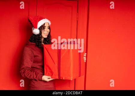 Junge Frau in Weihnachtsmann Hut hält großes Geschenk, steht in der Nähe der roten Wand. Glückliches Weibchen in Weihnachtsmütze mit Geschenkbox. Konzept der Ferien, präsentiert ein Stockfoto