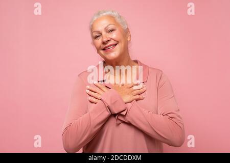 Ältere Frau bekommt immer wieder Komplimente. Entzückte glücklich charmante alte Dame mit weißen Haaren lächelt an der Kamera. Studio auf rosa Wand aufgenommen. Stockfoto