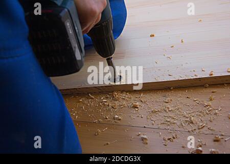 Ein Mann mit einem elektrischen Handbohrer für die Arbeit mit einem Holzbrett, Löcher und Türscharnier, Sägemehl überall Stockfoto