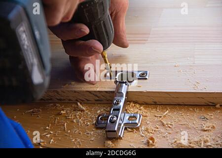 Ein Mann mit einem elektrischen Handbohrer für die Arbeit mit einem Holzbrett, Löcher und Türscharnier, Sägemehl überall Stockfoto