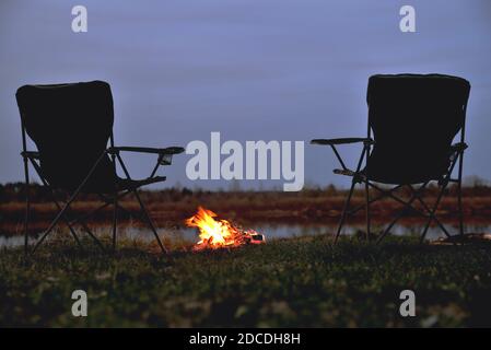 Ein Paar Touristenstühle, ein Klappstuhl, ein Lagerfeuer am Flussufer am späten Abend, Dämmerung im Herbst bei Sonnenuntergang, ein großes Lagerfeuer im Dunkeln, Stockfoto