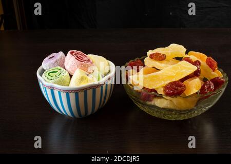 Türkische Freude an einer Porzellanvase und gelben kandierten Melonenscheiben, Ananasringen und Dogwood in einer Glasvase auf schwarzem Hintergrund. Stockfoto