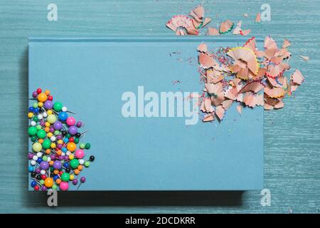 Blaues Hardcover-Buch mit Buntstiften, Späne und ein paar Daumenhacken auf einem hölzernen Schultisch. Stockfoto