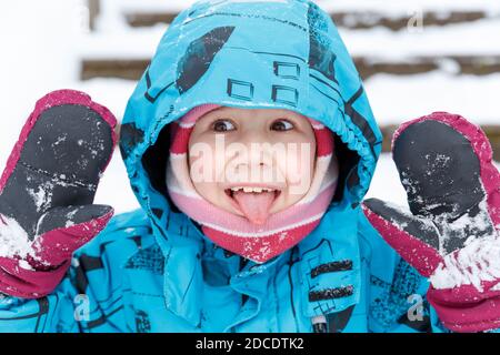 Nahaufnahme Porträt von fröhlichen kaukasischen Kind in Winterkleidung. Ein 6-7-jähriges Mädchen im Winter Overalls und Fäustlinge zeigt ihre Zunge und Handflächen. Wint Stockfoto