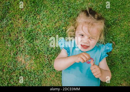 Zwei Jahre altes Mädchen liegt kopfüber und lächelt Der Rasen im Garten auf einer schönen Quelle Tag Stockfoto