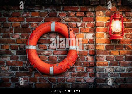 Alte, grungige und verwitterte Rettungsboje und rote Vintage beleuchtete Öllampe an rustikaler roter und brauner Ziegelwand. Moody Hintergrund oder Textur Stockfoto