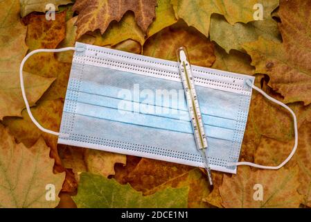 Blaue Gesichtsmaske und Glasthermometer zur Messung der Temperatur des menschlichen Körpers auf trockenen Herbstblättern. Stockfoto