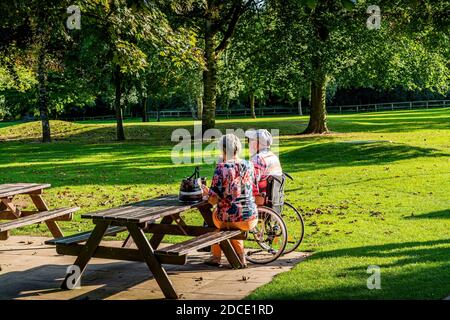 Warrington/Cheshire/UK 21/09/2020 Rentner, Frau, die sich um ihren behinderten Mann kümmert, der sich auf einer Parkbank niederlässt und sich in der Sonne entspannt Stockfoto