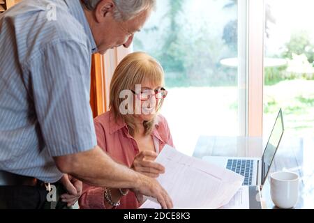 Eine ältere Frau und ein Mann lächeln und reden über Papiere. Die Frau ist ein Tisch mit einem Laptop vor ihr. Verwaltung Home-Konzept. Klare Lig Stockfoto