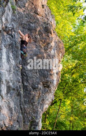 Glücklich schöne kaukasische Frau mit langen braunen Haaren aktiv Rock Klettern im Freien in einer schönen Landschaft zwischen grünen Bäumen und Klippen an einem sonnigen Tag Stockfoto