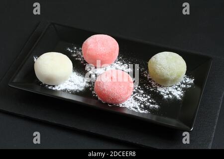 Vier Arten von japanischen Dessert-Mochi - Granatapfel mit Honig, grüner Matcha-Tee, Erdbeere, Kokosnuss auf einem schwarzen Teller auf einem schwarzen Tisch. Nahaufnahme Stockfoto