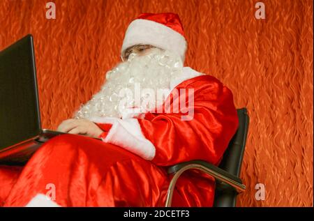 Der Weihnachtsmann sitzt in seinem Zimmer auf einem Stuhl mit einem Laptop in den Händen. Stockfoto