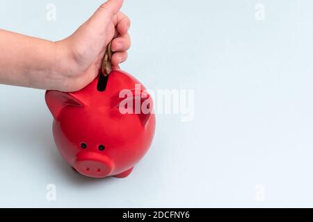 Nahaufnahme eines Kindes Hand legt mehrere Münzen in einem roten Sparschwein neben einem pastellfarbenen Hintergrund, Draufsicht, Kopierraum. Sparkonzept Stockfoto