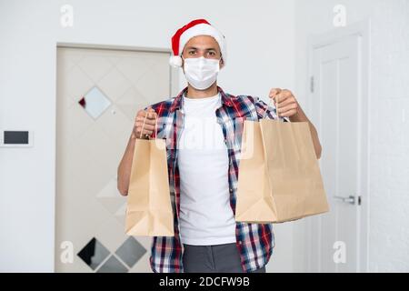 Männlicher Kurier in schützender medizinischer Maske und weihnachtsmann Hut hält Karton. Warenbestellung und Lieferung in Quarantäne. Stockfoto