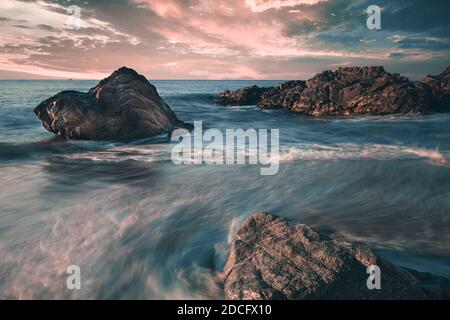 Seidiges Wasser und dramatischer Sonnenaufgang mit Felsen am Strand von Sant Pol de Mar, Katalonien, Spanien Stockfoto