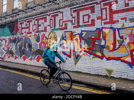 Man fährt Fahrrad auf der Grey Eagle Street, Shoreditch East London, mit lebendigen, bunten, künstlerischen Graffiti an den Wänden. Stockfoto