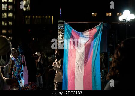 Menschen sammeln sich und halten Mahnwache am Transgender Tag der Erinnerung an verlorene Leben auf Washington Square. Die Menschen erinnern sich an das Leben von Transgender-Opfern von Gewalt und feierten das Leben derer, die leben. Die Menschen trugen meist weiße Kleider in Erinnerung an diejenigen, die starben. Sie setzten Rahmen mit Opferporträts und legten weiße Blumen und zündeten Kerzen an. Transgender-Flagge ist im Vordergrund als Turm des Freedom Tower of World Trade Center in Farben der Transgender-Flagge, die auf dem Hintergrund zu sehen ist beleuchtet gesehen. Stockfoto