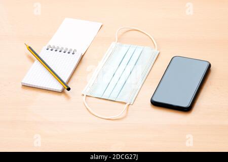 Auf einem Holztisch sind eine Gesichtsmaske, ein leerer Spiralnotepad mit Stift und ein Smartphone angeordnet. To-do-Liste Konzept während der Covid-19 Ausbruch. Stockfoto