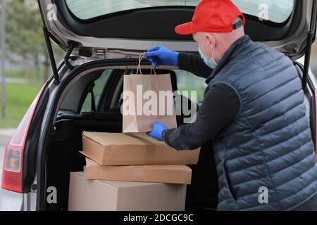 Delivery Mann in roter Kappe, Gesicht medizinische Maske und Handschuhe halten Papierbeutel in der Nähe des Autos im Freien. Service Coronavirus. Online-Shopping. Mock up Stockfoto