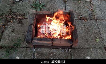 Mangal aus alten verbrannten Ziegeln auf Boden mit Zement quadratischen Blöcke bedeckt gebaut. Rustikale Outdoor-Szene. Lagerfeuer orange Flammen. Verkohlte Holztextur Stockfoto