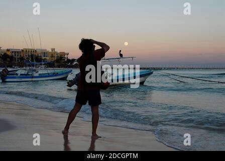 Ein unerkennbarer Fotograf am Strand bei Sonnenuntergang fotografiert die Landschaft. Im Hintergrund verankerten Boote, ein Pelikan und der Mond am Himmel Stockfoto