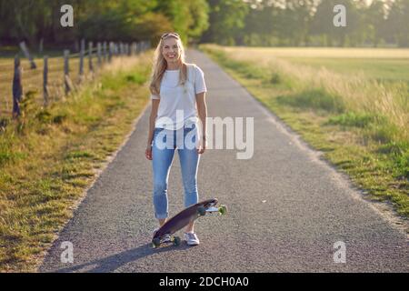 Attraktive athletische blonde Frau mit ihrem Skateboard von der Hintergrundbeleuchtung Abendsonne auf einer Landstraße zwischen Feldern lächelnd Die Kamera Stockfoto