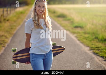 Porträt einer fit schöne Frau mittleren Alters mit einem aktiven Lebensstil lächeln und Blick auf die Kamera, während ein Longboard auf einer sonnigen Straße in der Stockfoto