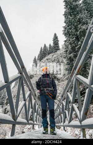 Bärtiger männlicher Reisender spaziert im Winter im Freien. Ein Kerl steht auf einer Brücke über einen Bergfluss. Wald in den Bergen nach einem Schneefall Stockfoto