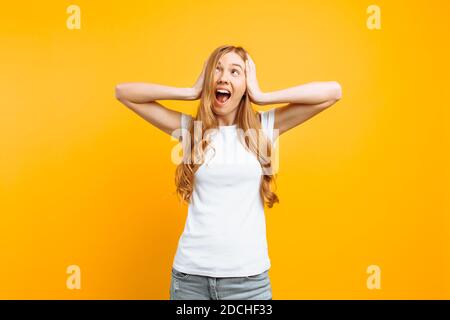 Porträt einer irritierten Frau in einem weißen T-Shirt, das ihre Ohren bedeckt und vor einem schrecklichen Geräusch schreit, auf einem gelben Hintergrund Stockfoto