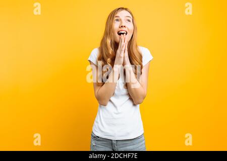 Junge fokussierte Mädchen bittet Gott, ihr zu helfen, hält die Hände in der Nähe ihres Gesichts, wünscht viel Glück, auf einem gelben Hintergrund Stockfoto