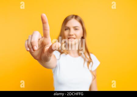 Frau in einem weißen T-Shirt, berührt einen imaginären Bildschirm mit einem Finger auf einem gelben Hintergrund Stockfoto