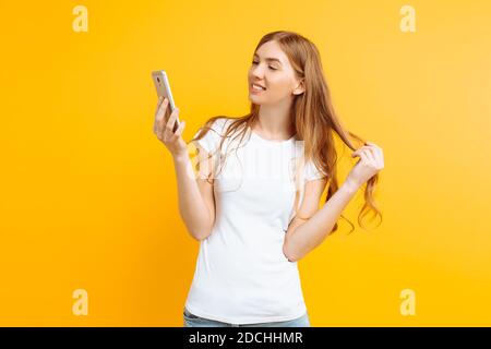 Schöne junge Frau in einem weißen T-Shirt, nutzt ein Handy, für Messaging, sieht auf dem Bildschirm eines Smartphones, auf einem gelben Hintergrund Stockfoto