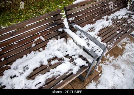 Braune Holzbank mit Schnee unter den Bäumen Herbst bedeckt Blätter Stockfoto