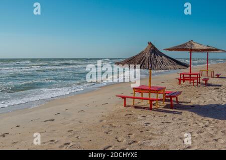 Sommerlandschaft: Tisch, Bänke und Sonnenschirm am Strand. Goldener Sand, stürmisches und winkendes Meer und blauer Himmel. Stockfoto