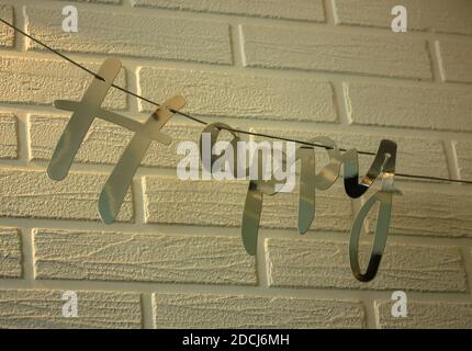 Das Wort Happy ist aus silbernem Papier vor einem weißen Backstein Wand Hintergrund. Festtagsgirlande aus Papier, Dekor für Geburtstagsfeier. Stockfoto