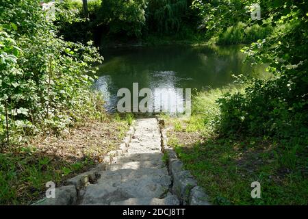 Treppen aus Steinblöcken gehen bergab zum Fluss. Das Becken ist unter Vegetation versteckt und teilweise von ihm bedeckt. Der Wasserfluss ist langsam. Stockfoto