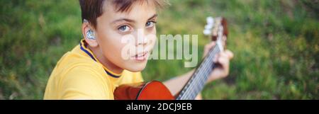 Schwer zu hören preteen junge spielen Gitarre im Freien. Kind mit Hörgeräten in den Ohren, das im Park Musik spielt und singt. Hobby Kunst Aktivität Stockfoto