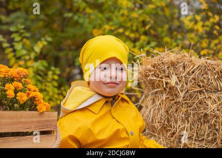 Niedliche schöne Pressoler Junge in einer orangen Hose, Regenmantel, Hut, Gummistiefel in der Nähe eines Haufen Heu. Gemütlichkeit, Herbst. Stockfoto