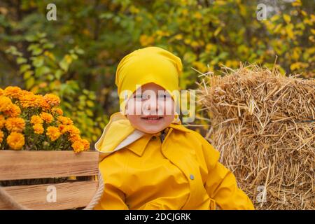 Niedliche schöne Pressoler Junge in einer orangen Hose, Regenmantel, Hut, Gummistiefel in der Nähe eines Haufen Heu. Gemütlichkeit, Herbst. Stockfoto